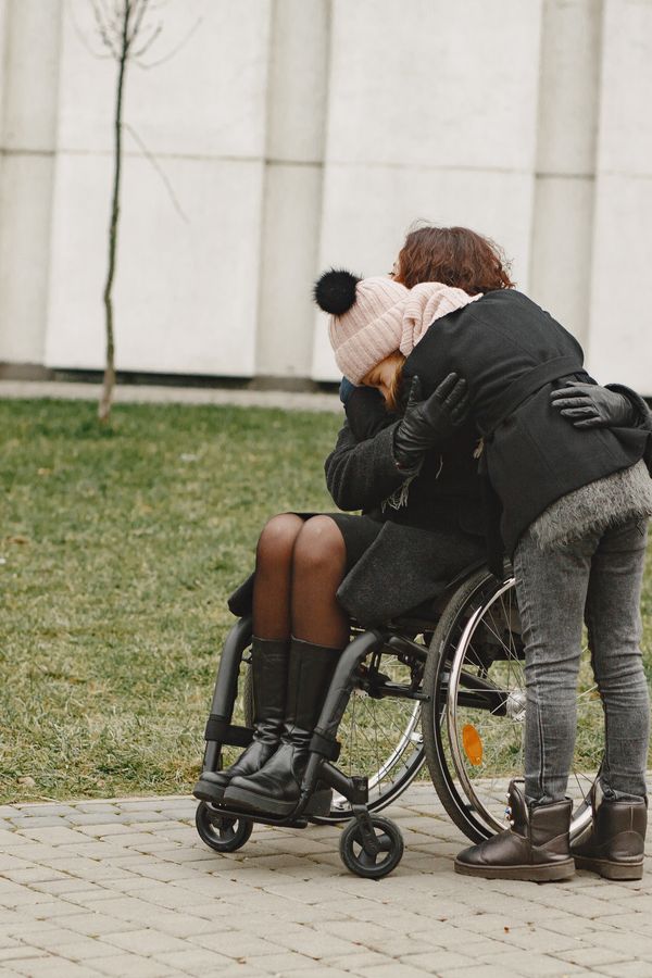 Friends embracing warmly outdoors, one using a wheelchair